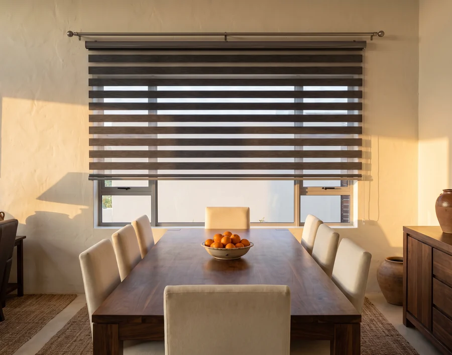 Charcoal day-night blind face-fixed above a Woodhill dining-room window, its sheer and solid bands letting morning light fall in stripes across a walnut table